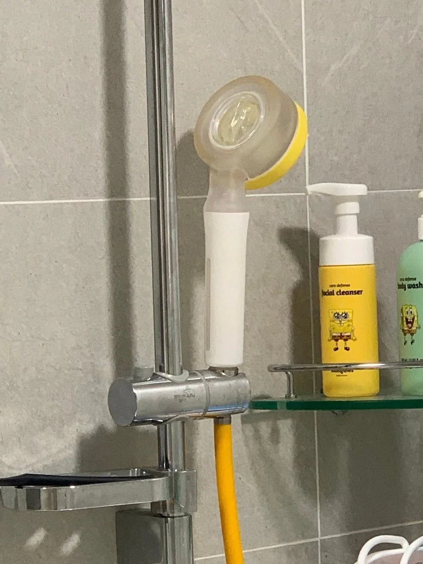 Close-up of a bathroom shower area with a yellow and white facial cleansing brush, a yellow Mr. Clean themed facial cleanser bottle, and a green SpongeBob themed body wash on a glass shelf.