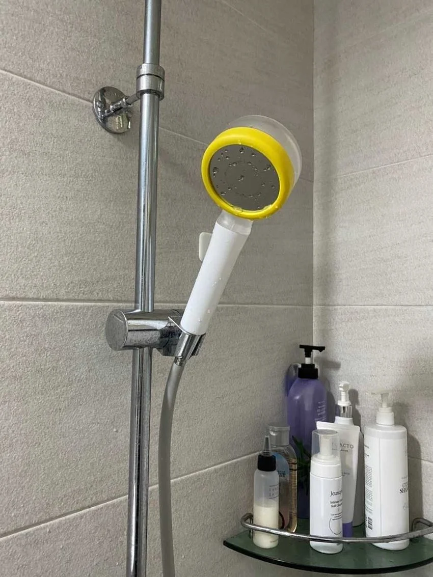 A shower head with a yellow and gray shower filter attached, mounted on a tiled bathroom wall. Several bottles of shower products are on a glass shelf below.