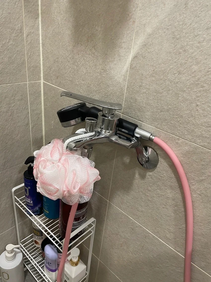Bathroom shower faucet with pink hose, mounted on beige tiled wall, with a pink bath sponge hanging from the faucet. Shelves below hold bottles of shampoo, conditioner, body wash, and other toiletries.