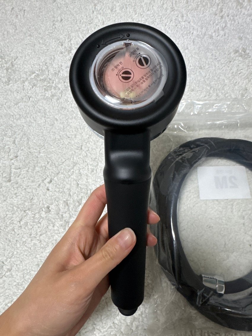 A person holding a black handheld device with a transparent circular opening at the top, showing a metal part inside, next to a coiled black cable, on a light-colored textured surface.