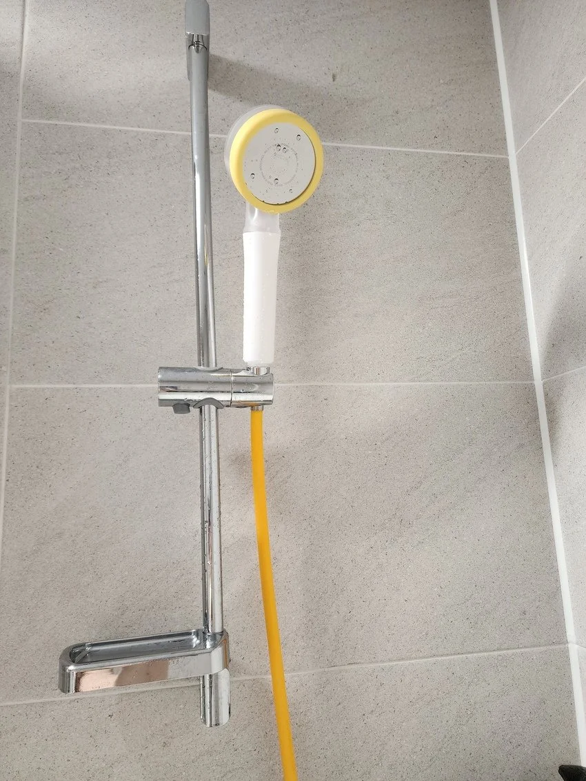 A showerhead with a yellow hose connected to a chrome shower pipe mounted on a beige tiled wall.