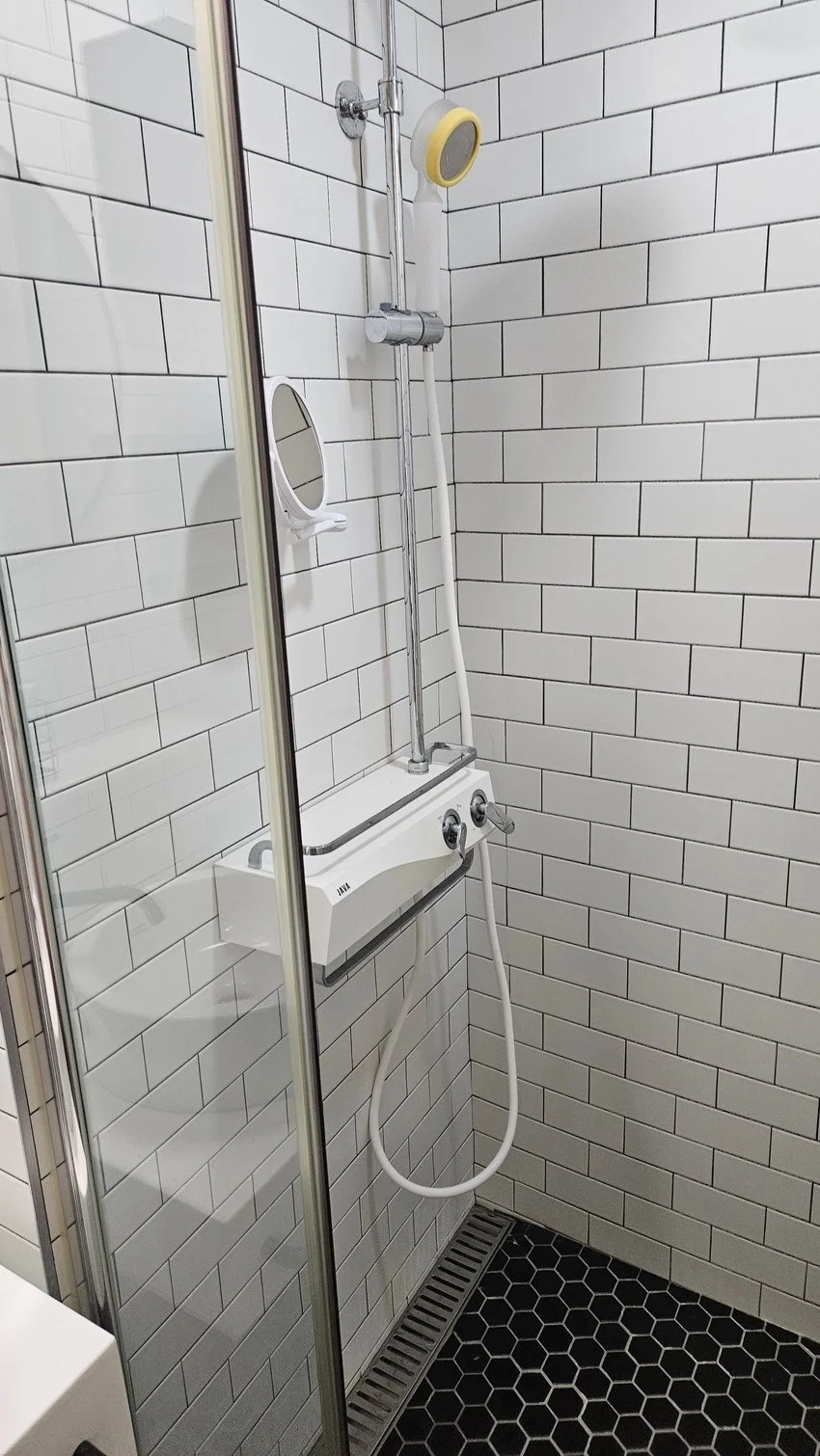 Shower area with white subway tiles, a showerhead, a hand-held shower, a small mirror, and a drain on black hexagonal tile floor.