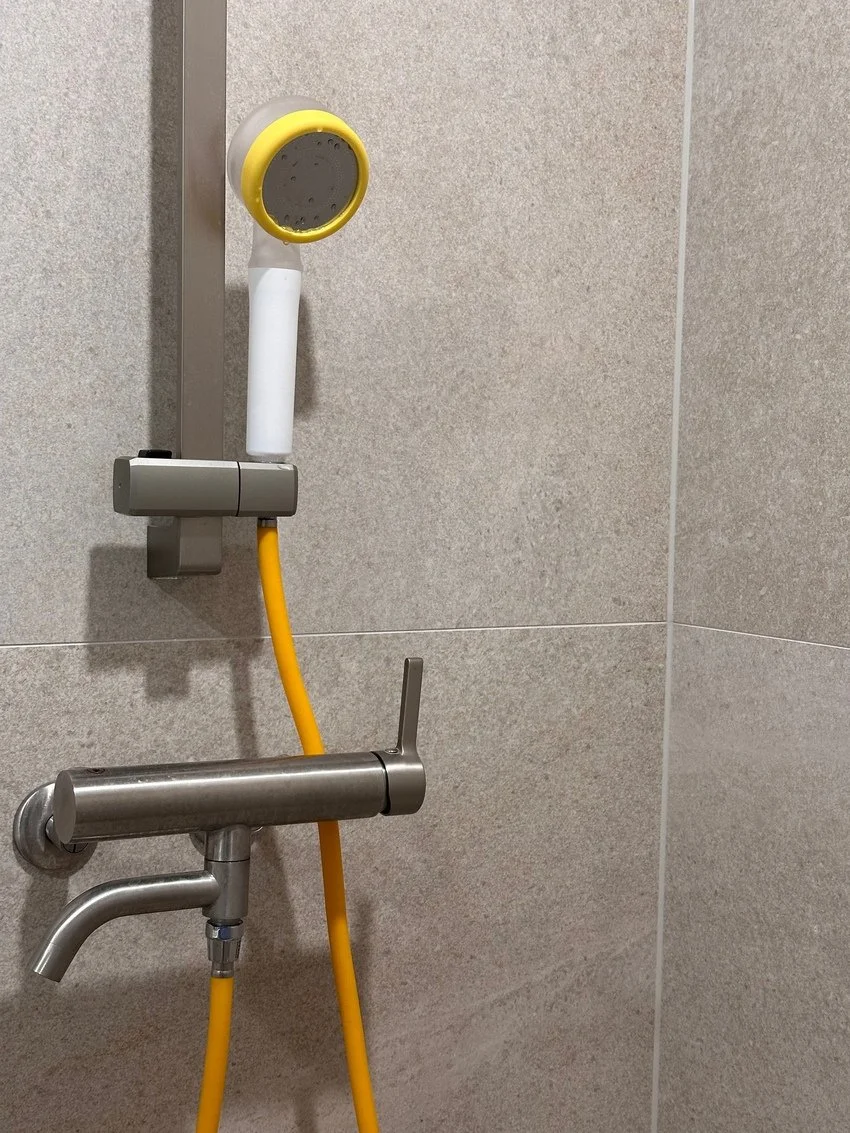 A shower with a yellow handheld showerhead, a gray faucet, and yellow hose in a bathroom with beige tiled walls.