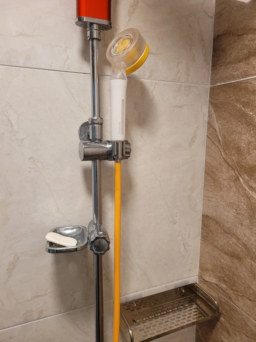 Showerhead, soap dish with soap, and yellow hose mounted on a tiled bathroom wall.
