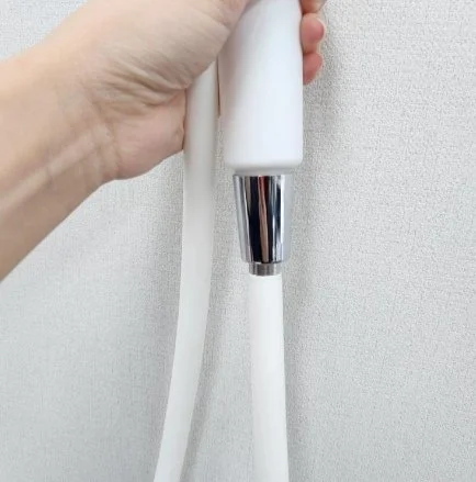 Hand holding a white showerhead attached to a hose against a white textured wall.