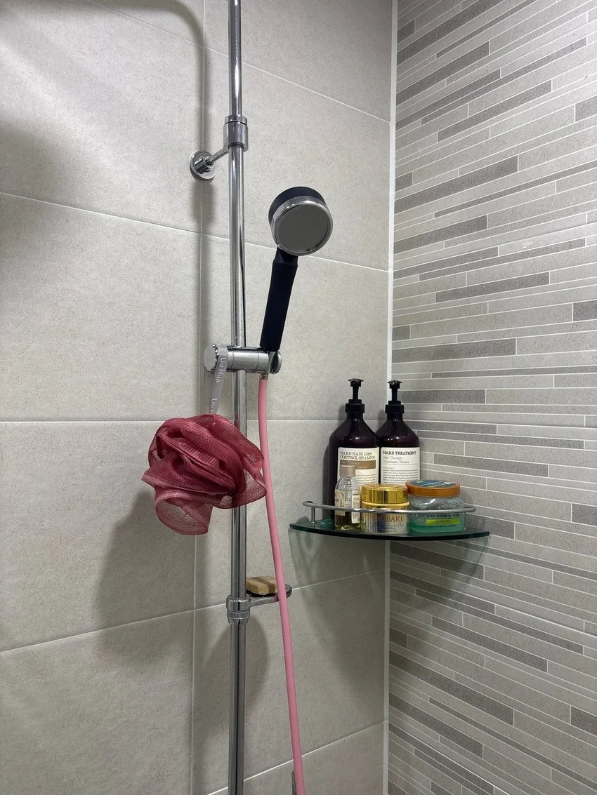 Shower area with handheld showerhead, shower gel, shampoo bottles, and small containers on a glass shelf.