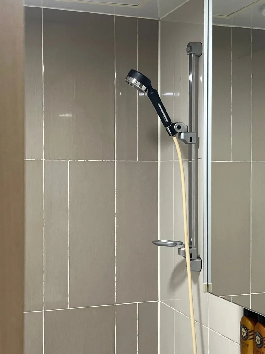 Close-up of a shower head and sliding shower door in a bathroom with beige tiled walls.
