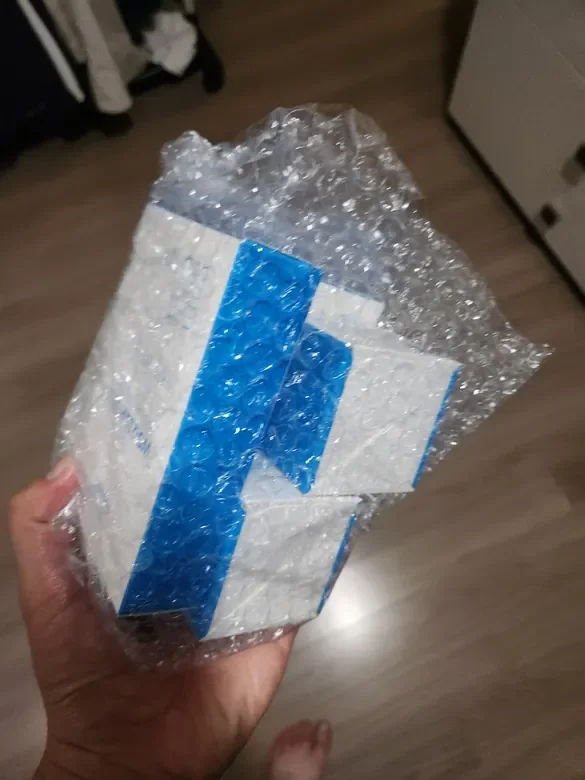 A hand holding a package wrapped in bubble wrap inside a room with wooden flooring and some furniture.