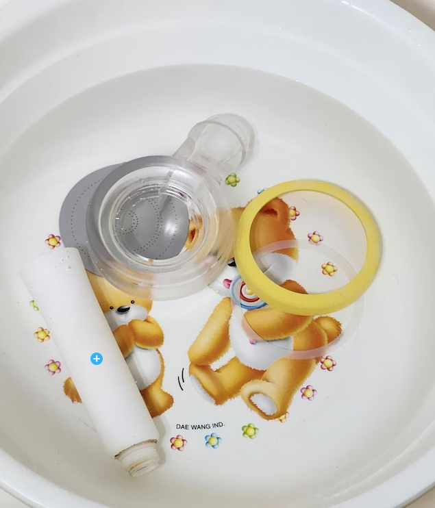 Inside a white basin with toy bears, a roll of paper, a transparent baby bottle, and a yellow ring. The basin has small flower illustrations and the text "DAE WANG IND."