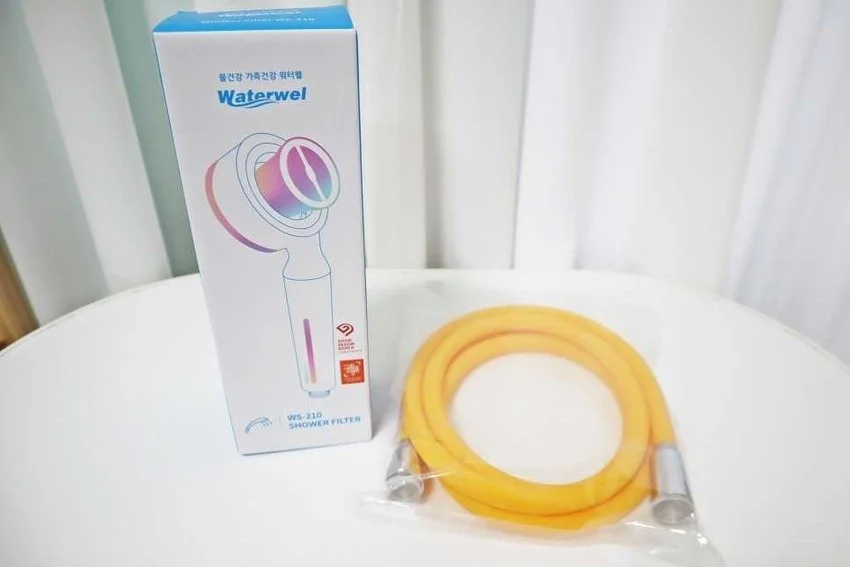 Waterwel shower filter in white box with blue text and a yellow shower hose in plastic packaging on a white surface.