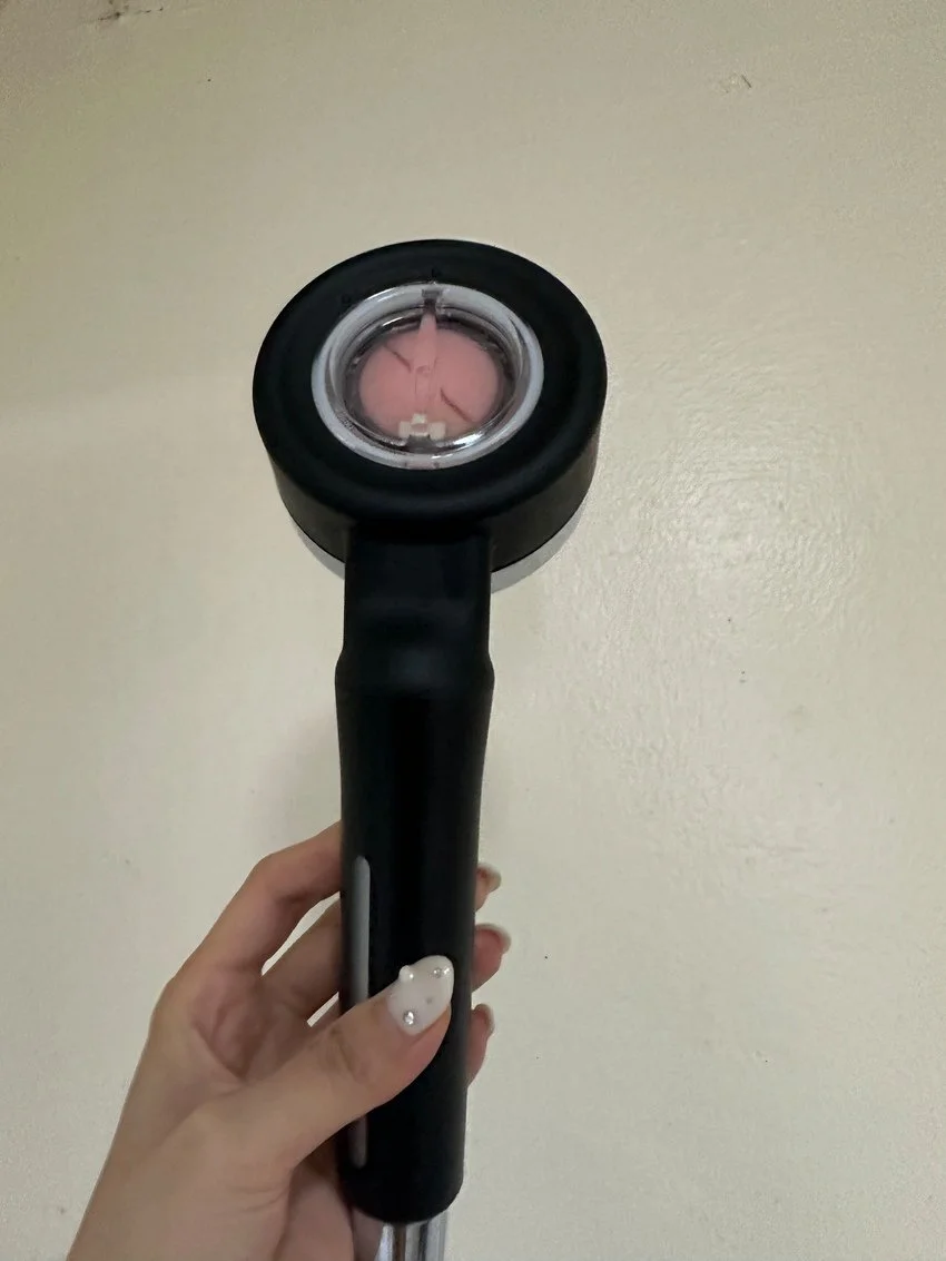 Person holding a black handheld device with a pink object inside, against a plain off-white wall background.