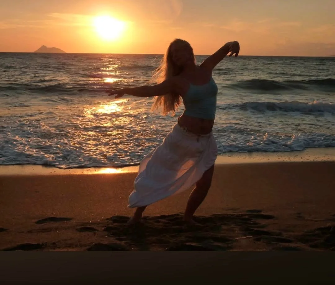 Eine Frau tanzt am Strand bei Sonnenuntergang, mit den Armen ausgestreckt und im Sand stehend.