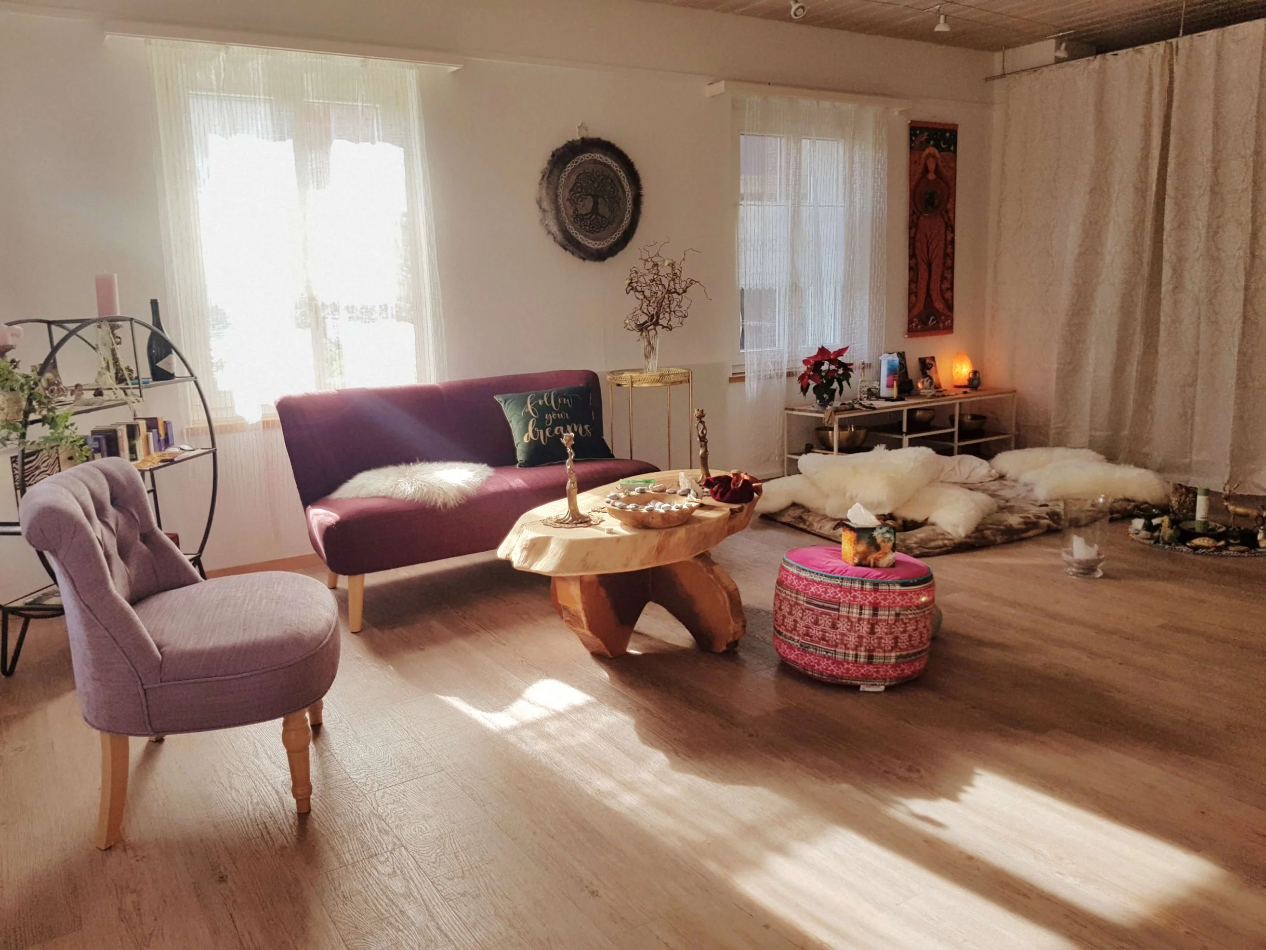 Ein Wohnzimmer mit großen Fenstern, einer rot-violettfarbenen Couch, einem runden Holztisch, einem bunten Sitzhocker, Dekorationsgegenständen, Wandbildern und einem Fußboden aus Holz. Es wirkt hell und gemütlich.