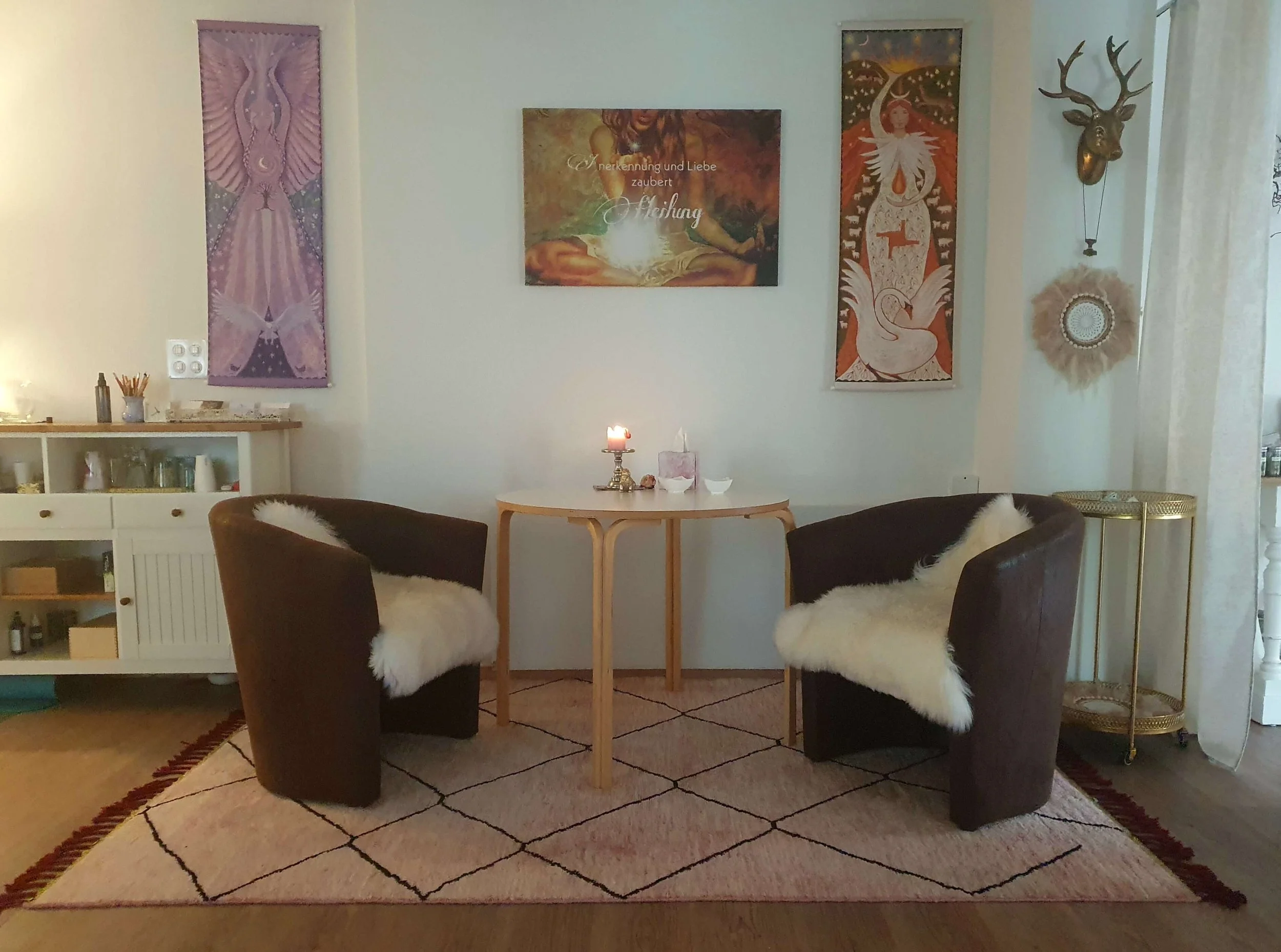 Ein Wohnzimmer mit drei Wandbehängen, einer Kerze, einer Schale und zwei Stühlen mit kuscheligen Überwürfen, einem Teppich und einem sideboard darunter.