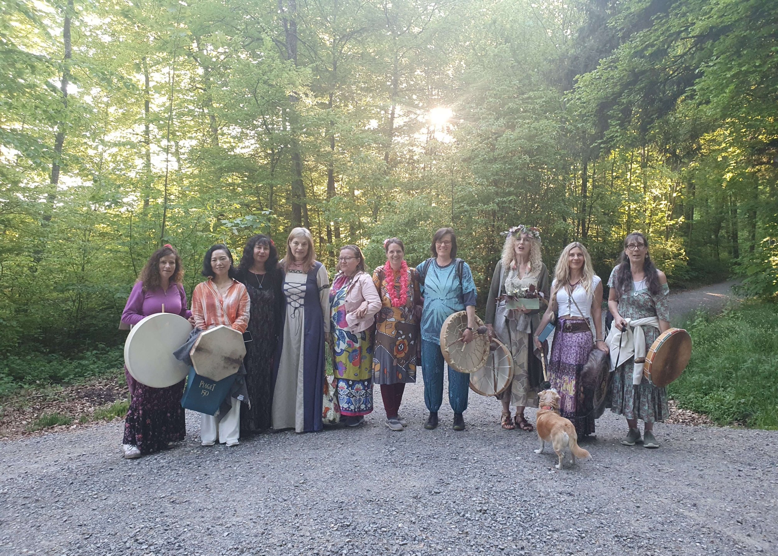 Eine Gruppe von Frauen steht im Wald bei Sonnenuntergang, einige tragen bunte Kleider und halten Musikinstrumente, darunter Trommeln und Rasseln, während Hunde neben ihnen sitzen.