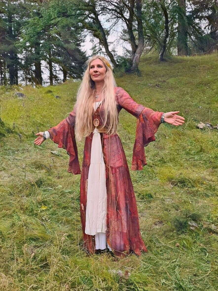 Eine Frau mit langen blonden Haaren steht im Grünen, trägt ein langes, bunte Kleid mit weiten Ärmeln und hält die Arme offen in einer Naturumgebung mit Bäumen und Gras.