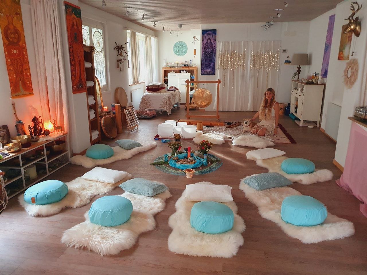 Ein Raum mit Sitzkissen und Fellen auf dem Boden, einer Frau mit Hund, einer zentralen Yoga- oder Meditationsstation, bunten Wanddekorationen und einer ruhigen, meditativ angehauchten Atmosphäre.