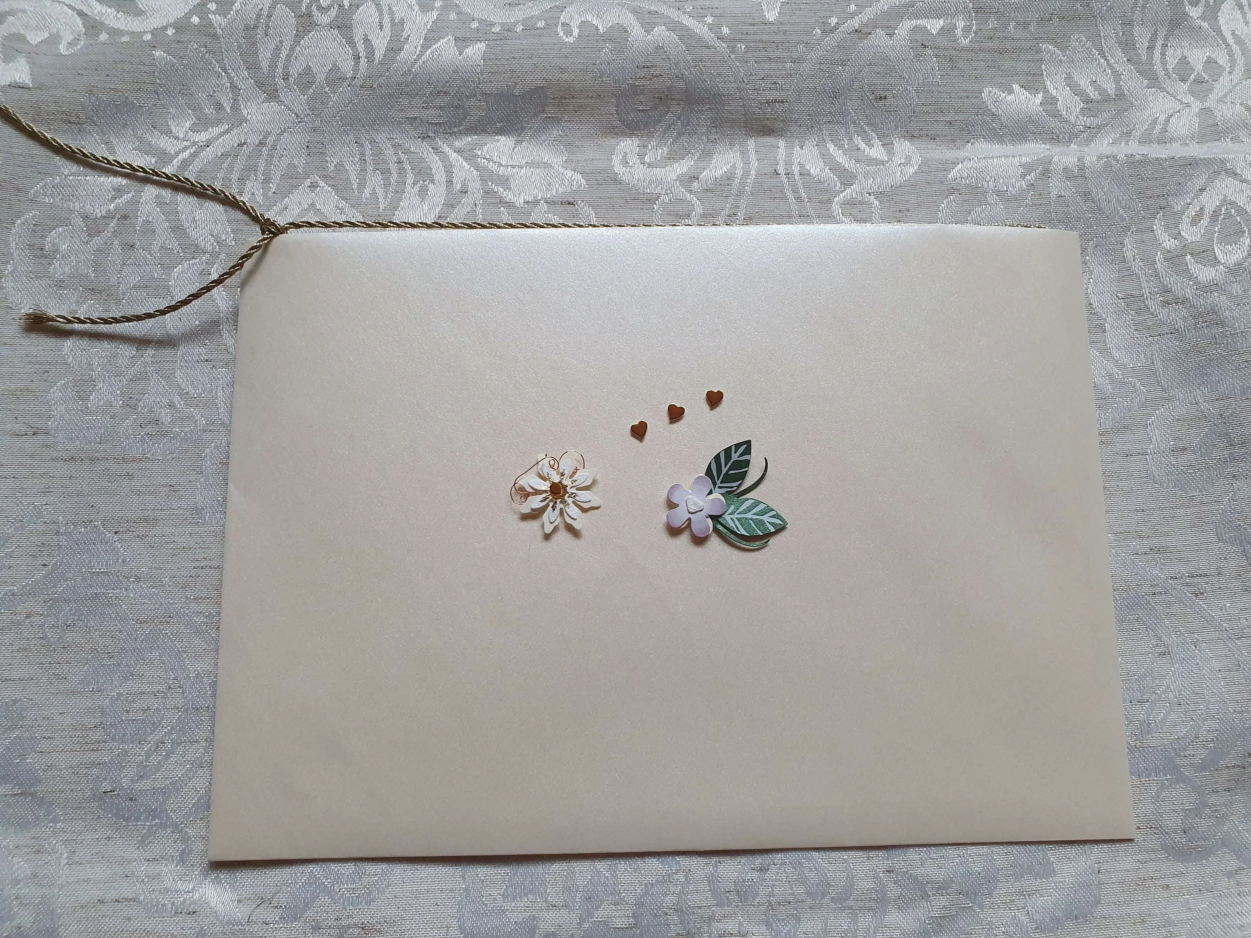 Weiße Karte mit kleinen roten Herzchen, weißen Blumen und grünen Blättern, auf einem silbernen, glänzenden Tischläufer
