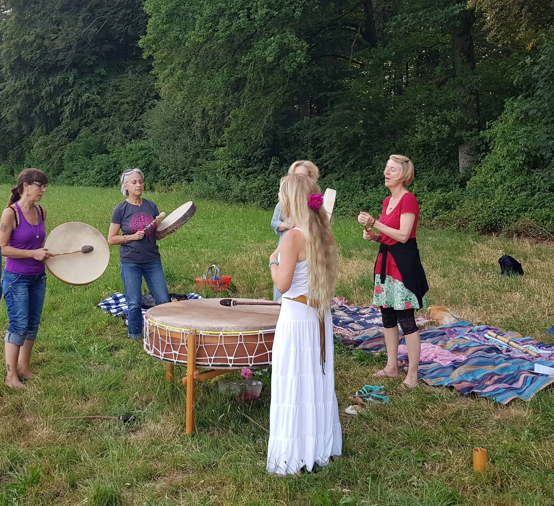 Eine Gruppe von Menschen, die im Freien in einer grünen Wiese stehen und sich an einer Zeremonie oder einem Ritual beteiligen, mit Musikinstrumenten wie Trommeln.