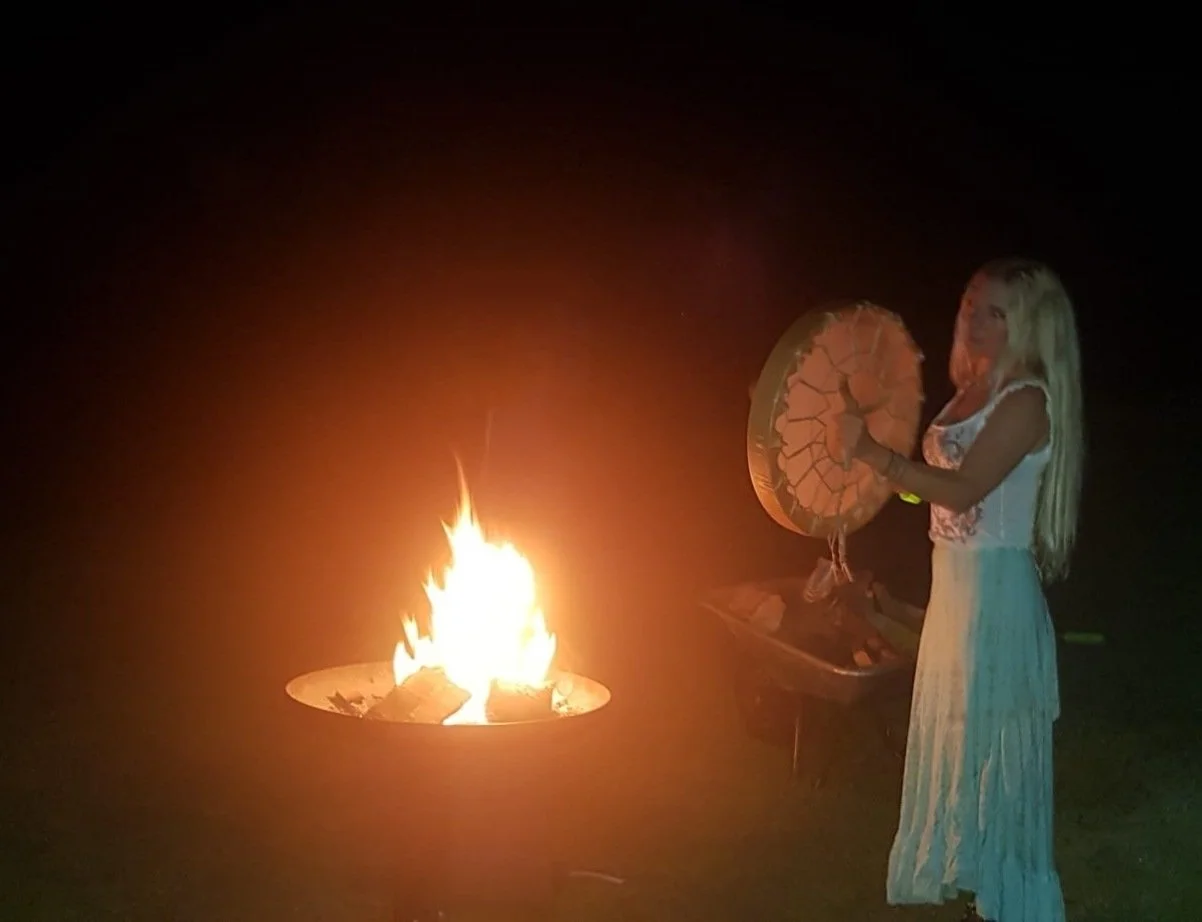 Junge Frau mit langem blonden Haar in weißem Kleid steht nachts neben einem Lagerfeuer und hält eine brennende Geoden-ähnliche Kugel in der Hand.