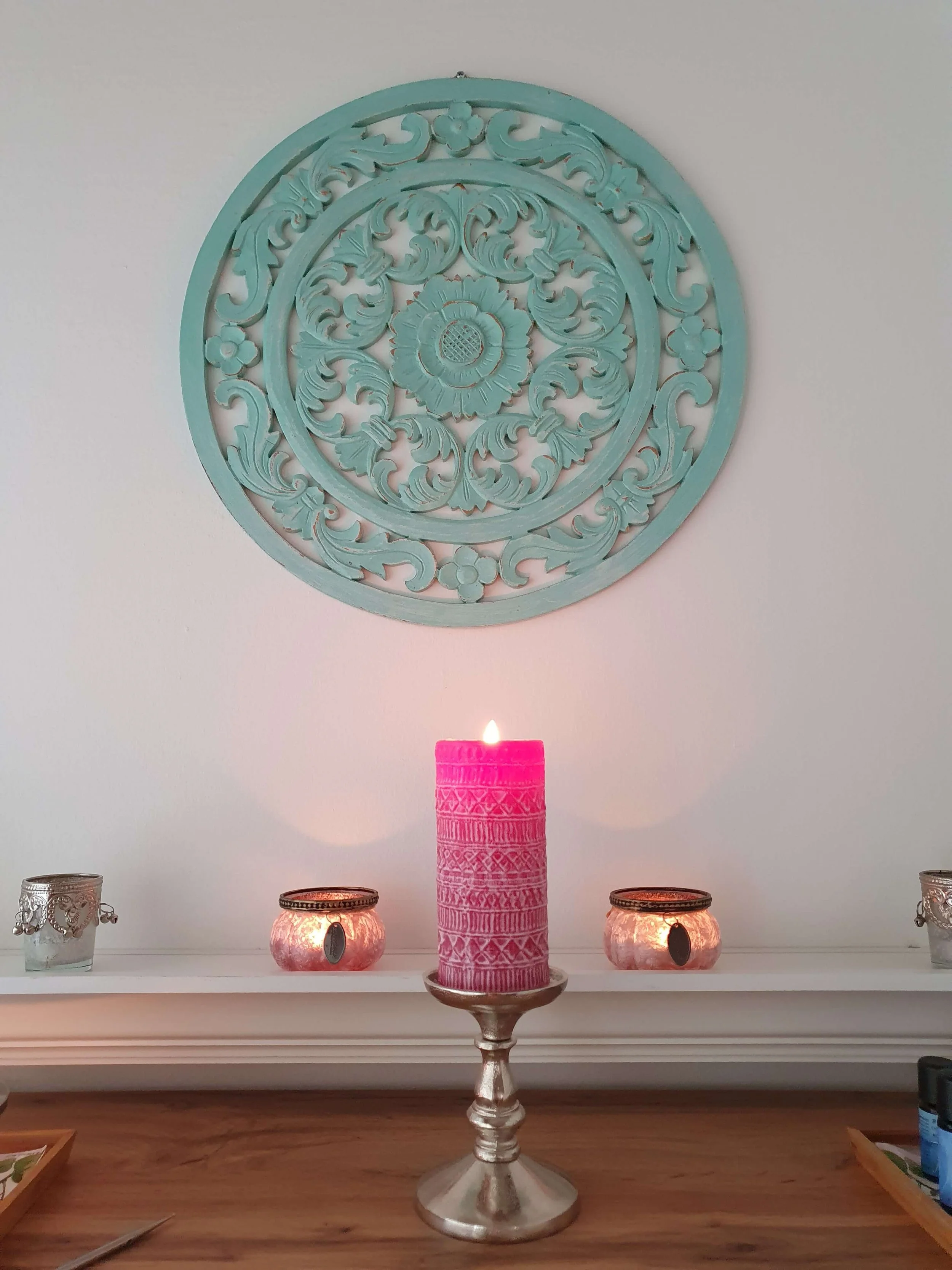 Ein geschnitztes, kreisförmiges Wandkunstwerk in Türkis hängt an einer weißen Wand. Darunter steht ein Kerzenständer mit einer großen pinken Kerze. Auf dem Tisch sind vier kleinere Laternen in Kupfer- und Silberfarben, sowie einige andere dekorative Gegenstände.