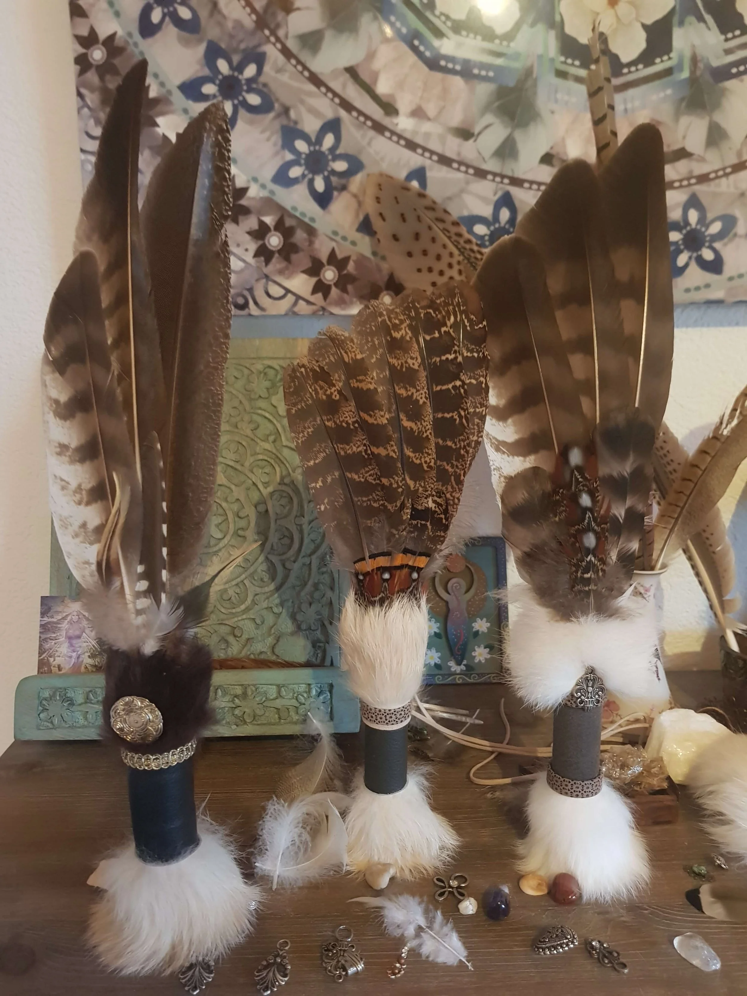 Verschiedene Federn in Braun-, Schwarz-, Weiß- und Beigetönen, einige mit Mustern, stehen auf schwarzen Ständen. Darunter liegen Schmuckstücke und kleine Ornamente auf einem Holztisch.