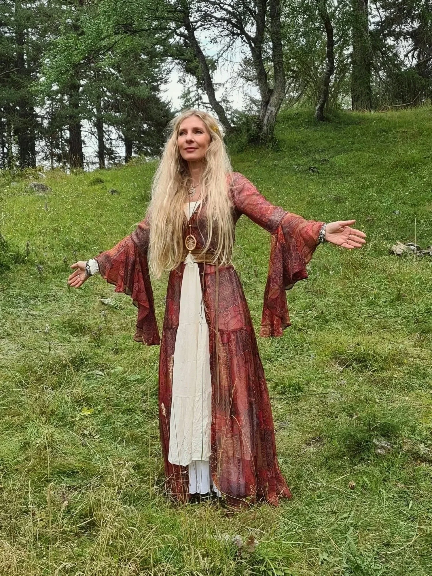 Eine Frau mit lang blondem Haar in einem langen, burgunderfarbenen Bohemian-Kleid steht vor Bäumen in einem Wald. Sie zeigt mit den Armen nach oben und zur Seite, wirkt meditativ oder spirituell.