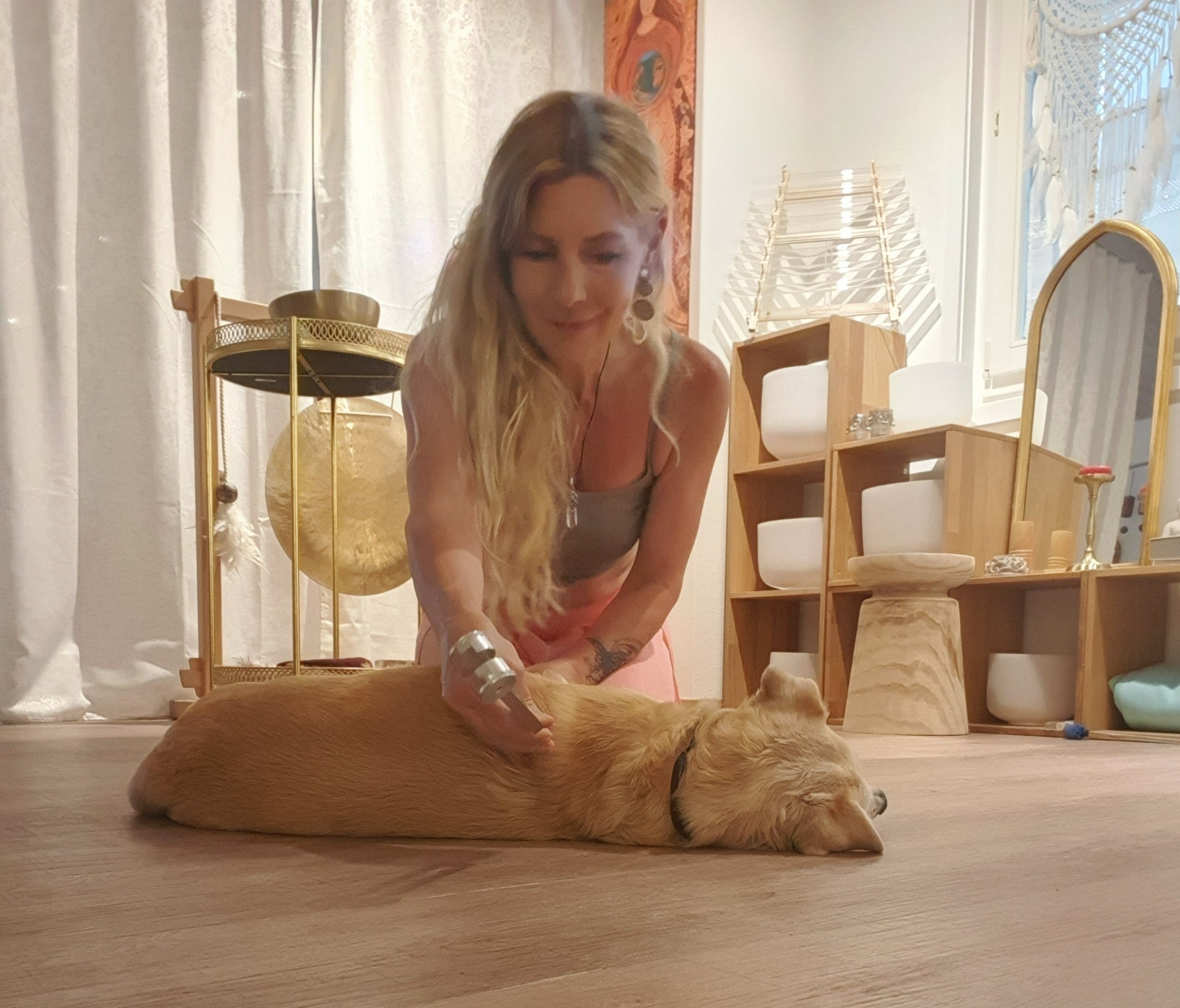 Eine Frau mit langen blonden Haaren kniet auf dem Boden neben einem liegenden Hund. Im Hintergrund stehen weiße Vorhänge, ein dekorativer Globus, Regale mit verschiedenen Gegenständen und ein Spiegel.