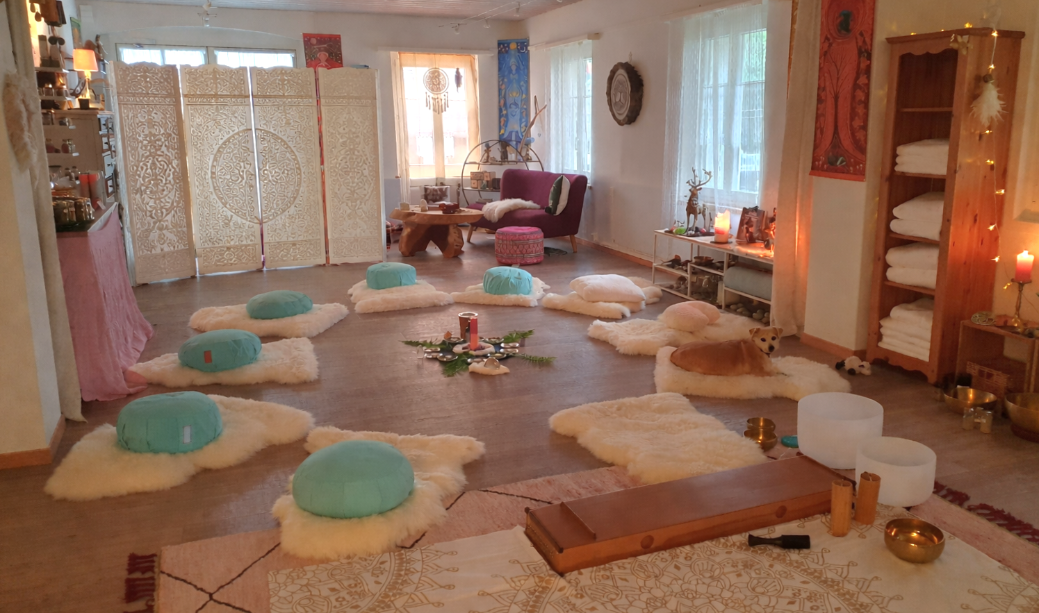 Ein gemütlich eingerichteter Raum mit Meditationselementen, Wolken- und Kissen-Elementen, einer deko mit Kerzen, einem Hund auf einem Futterteppich, sowie dekorativen Gegenständen und Handtüchern. Es wirkt wie ein Raum für Meditation, Yoga oder Rituale.