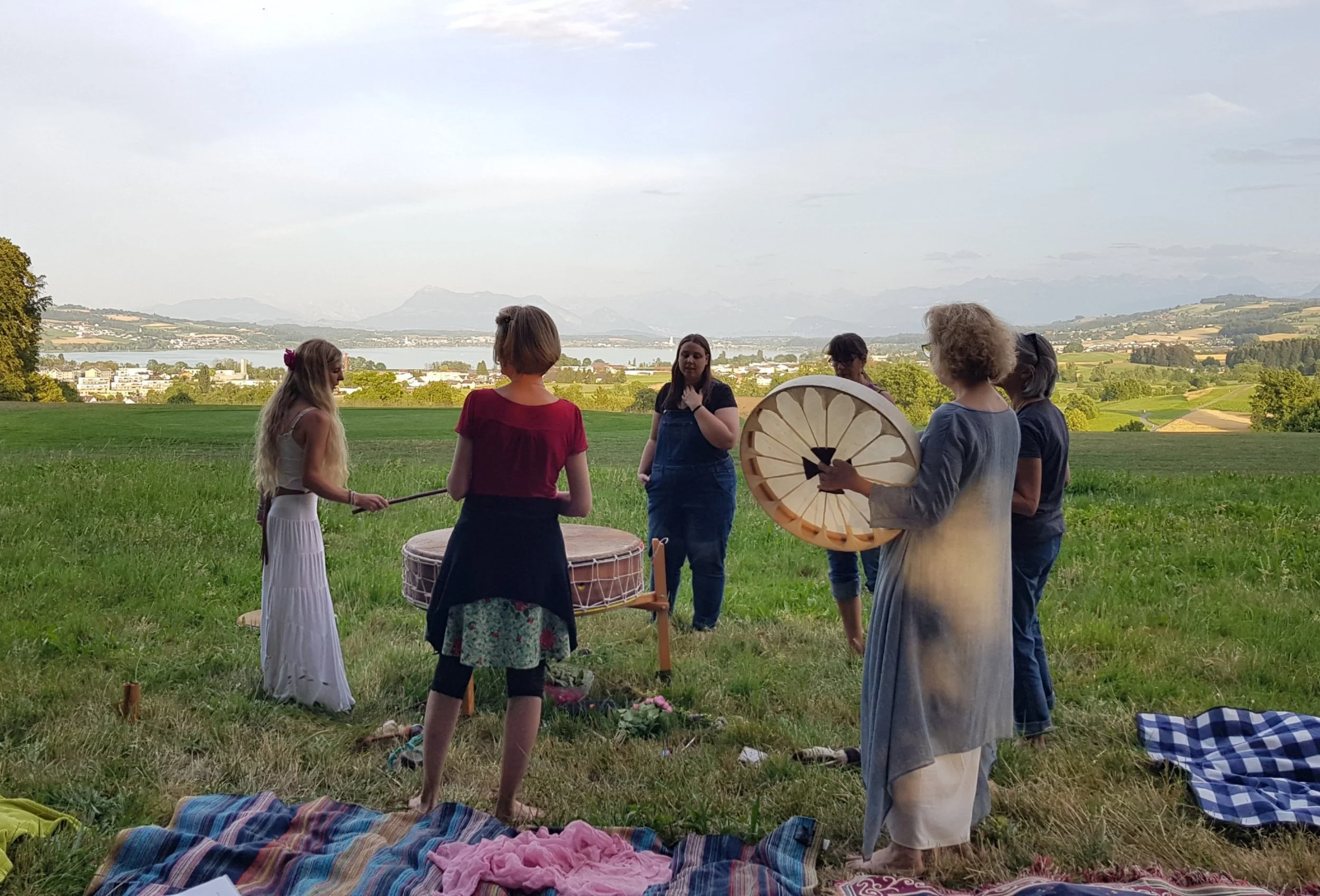 Gruppe von Frauen und Mädchen, die einen rhythmischen Tanz oder eine Musiksession im Grünen vor einer malerischen Landschaft mit See und Bergen durchführen.