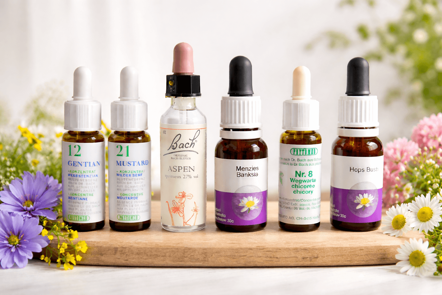 Verschiedene Fläschchen mit homöopathischen Mitteln, umgeben von Blumen auf einem Holzbrett.