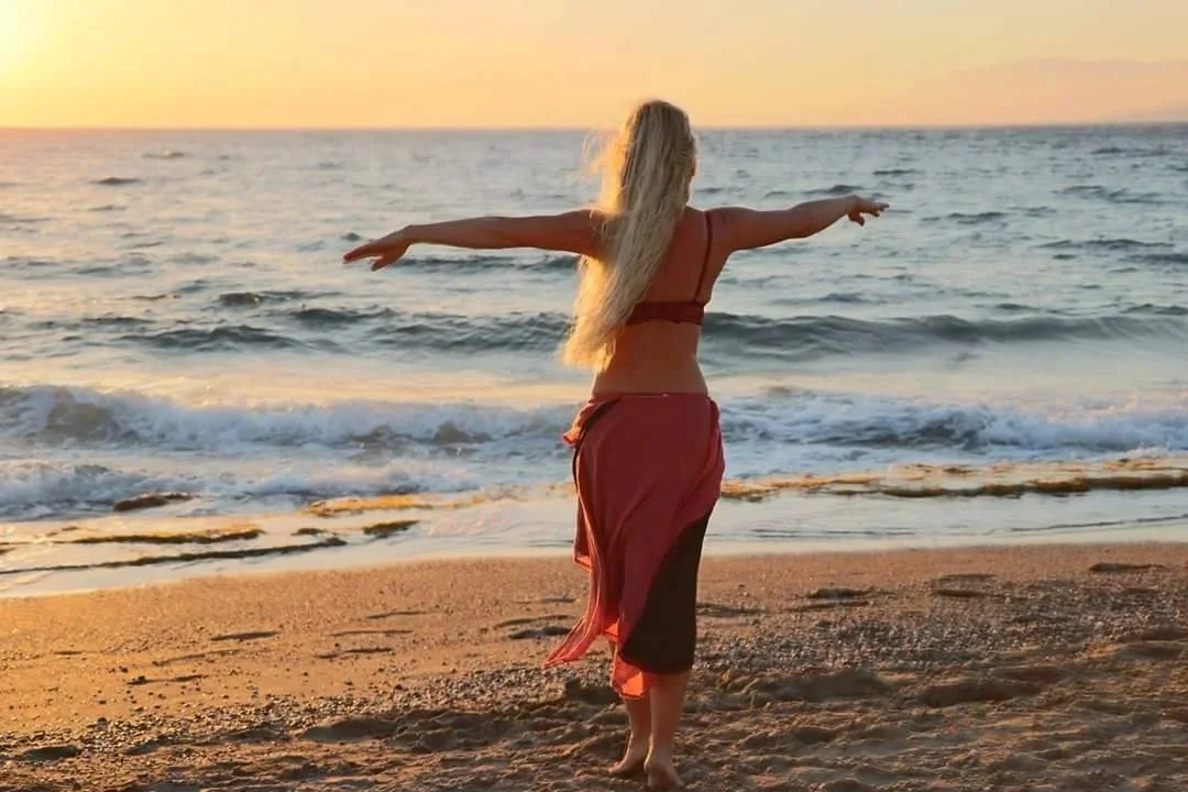 Frau am Strand bei Sonnenuntergang mit ausgestreckten Armen, Blick auf das Meer.