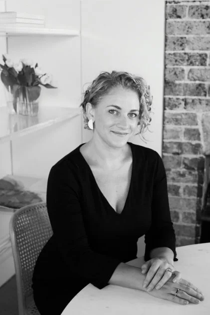 A woman with curly hair sitting at a table, smiling, indoor setting with a brick wall and a shelf with flowers in the background.