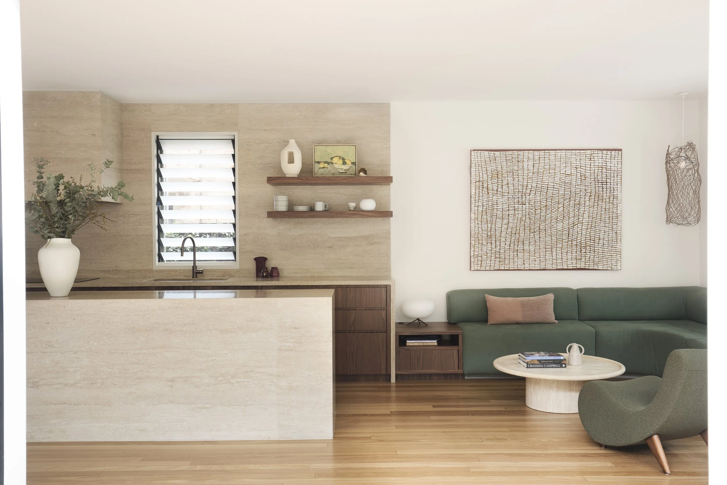 A living room with a green sofa, a round coffee table with books, a gray armchair, a wooden side table, wall art, and a large woven wall hanging. The room features a wood floor and white walls.