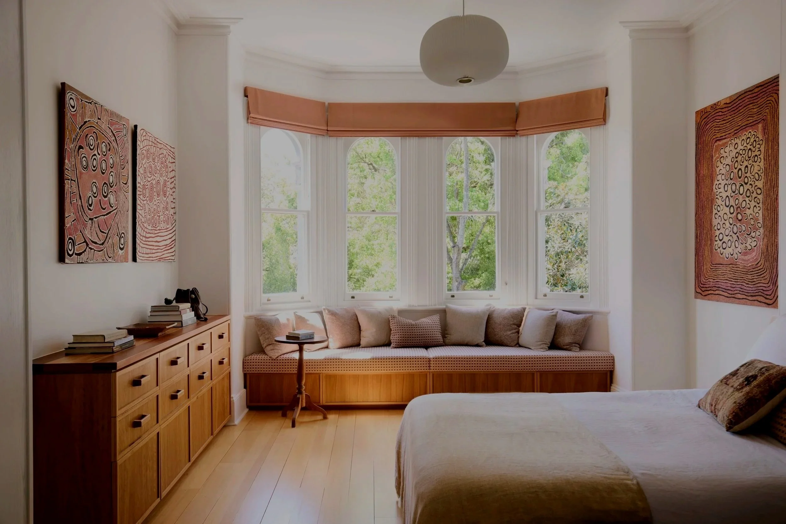 Living room with large bay window, beige sofa with cushions, wooden sideboard with books, and abstract artwork on the walls.