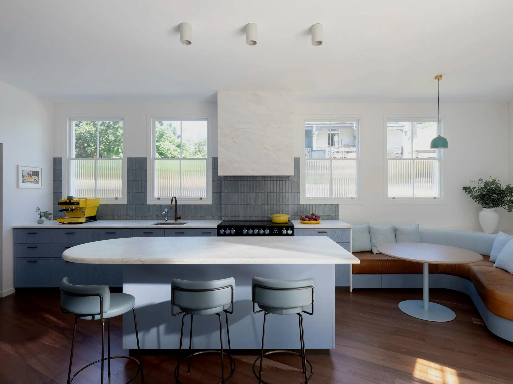 Modern kitchen with white walls, multiple windows, a marble island, and a built-in oven. There are bar stools around the island and a corner bench with cushions. A chandelier hangs above, with a plant visible on the right.