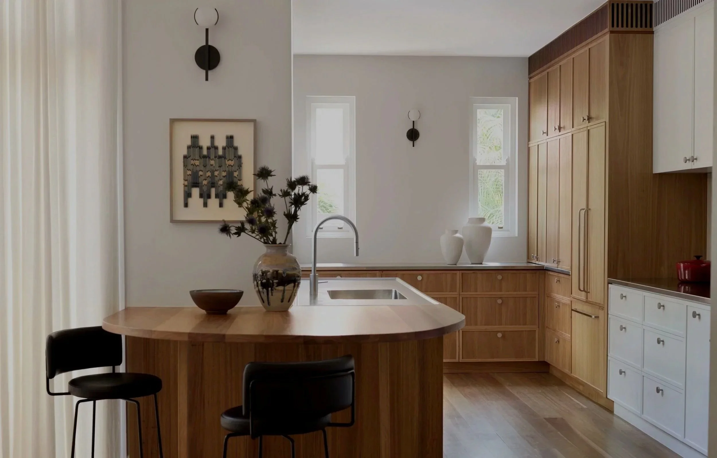 Modern kitchen with wooden cabinets, white walls, two white vases on window sill, black wall-mounted light fixtures, a round wooden dining table with black chairs, vase with flowers on the table, and windows allowing natural light in.