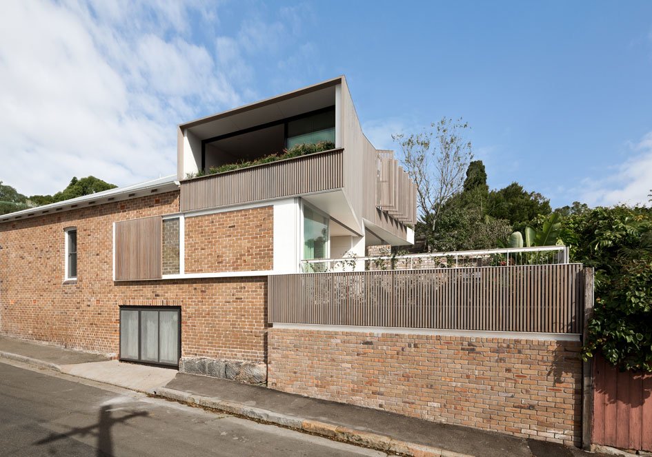 Modern multi-level house with brick and wood exterior, large balcony, greenery, and a clear blue sky.