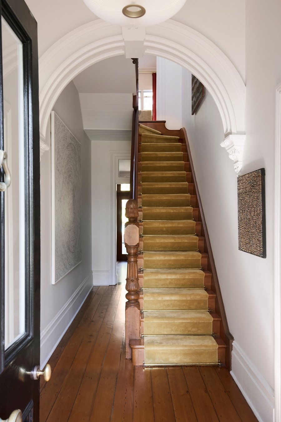 View of an interior staircase with a wooden handrail, beige carpet runner, and white walls, seen through an arched doorway in a house.