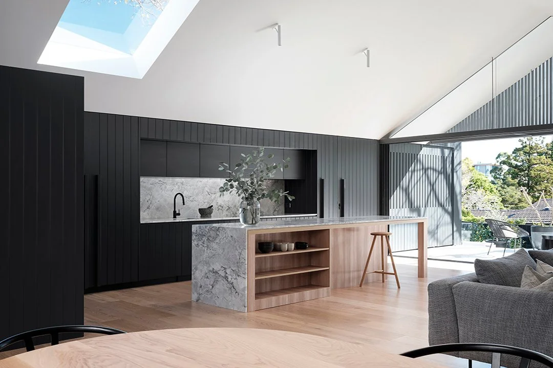 Modern open-plan kitchen with black cabinets, marble island with a wooden extension, gray sofa, wooden dining table, and large sliding glass doors leading to outdoor patio with trees.
