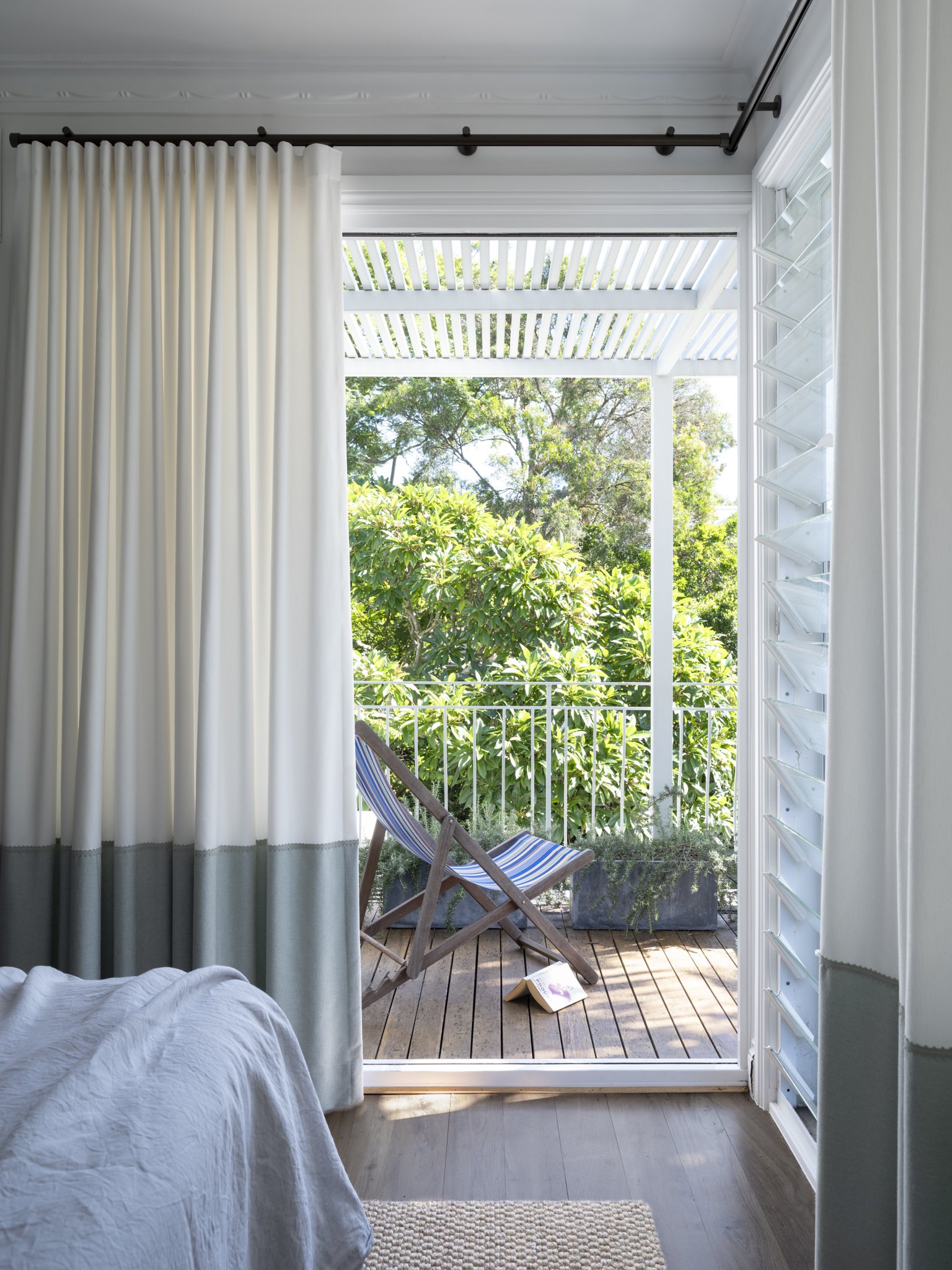 A cozy bedroom with a sliding glass door opening to a small balcony with greenery outside. The room has white curtains, a white bed, and a striped deck chair on the balcony, with a book on the wooden floor.