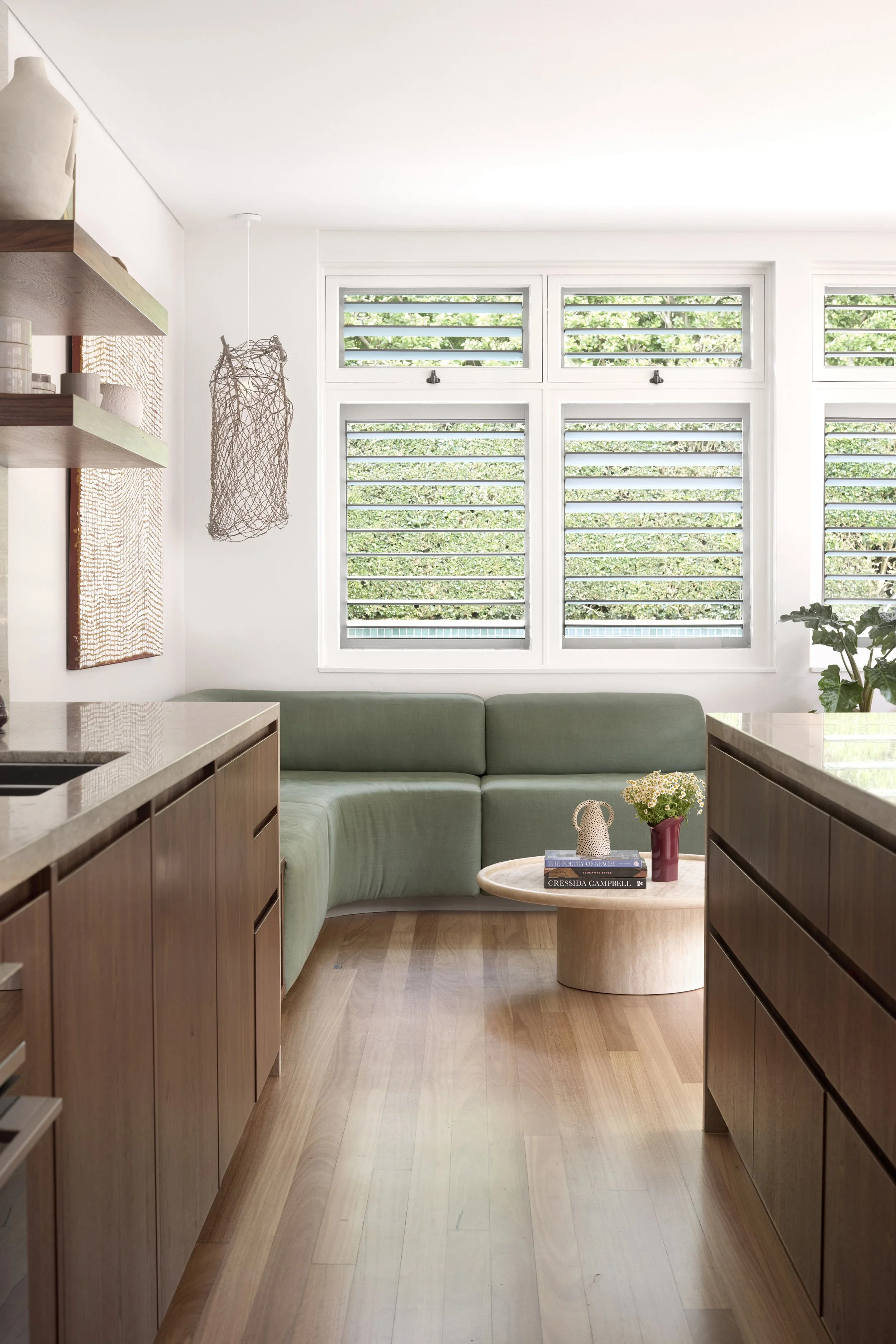 A modern living room with a built-in green sofa, a small oval coffee table with books and a flower vase, large windows with green trees outside, and wooden cabinets along the sides.