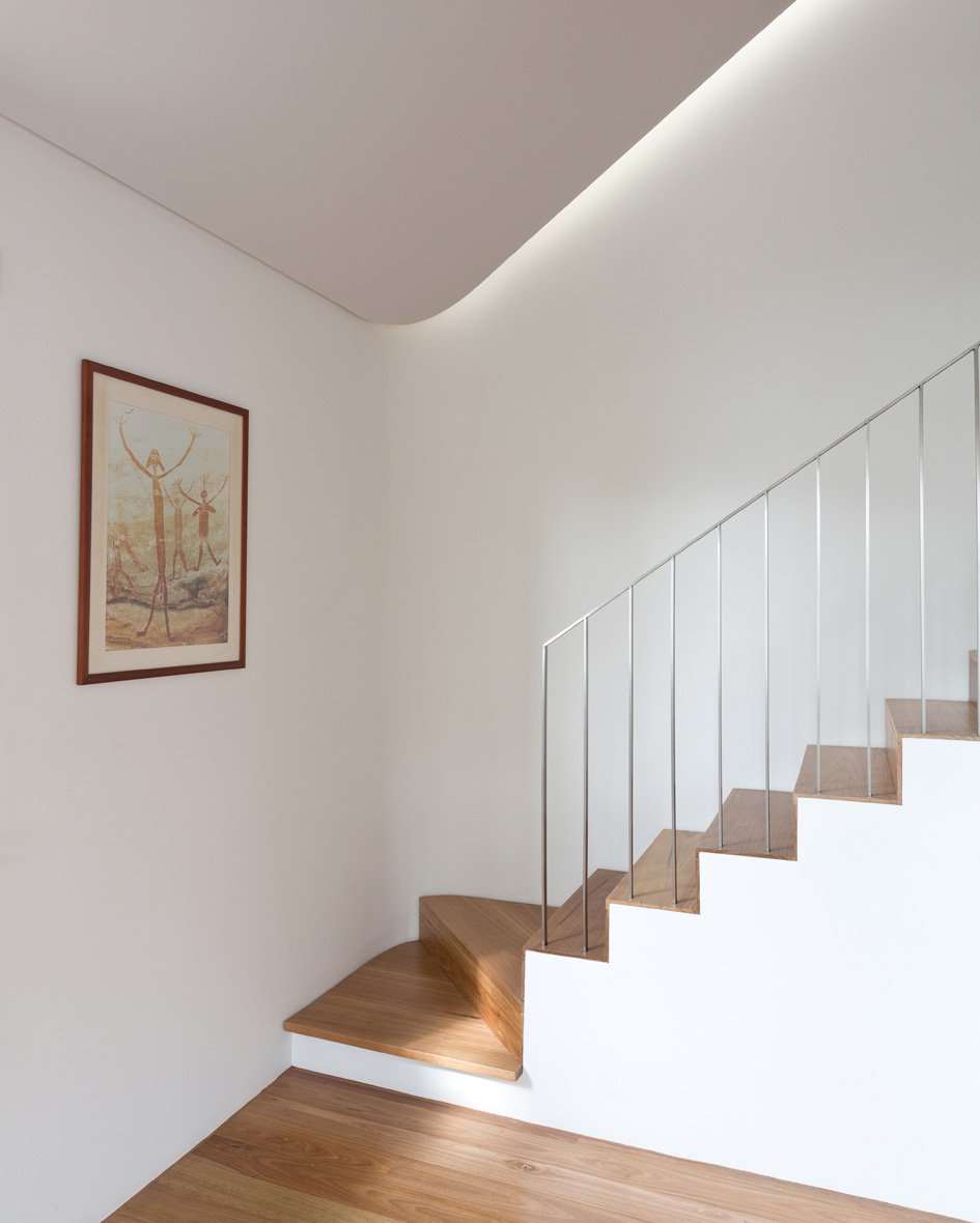 Interior of a modern home with a wooden staircase, a white wall, and a framed artwork depicting human figures with arms raised.