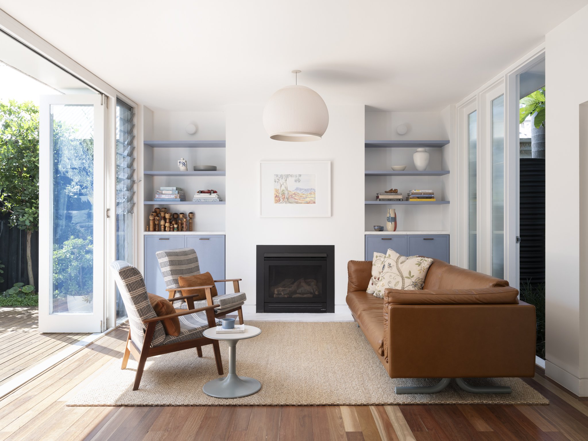 Living room with open door leading to outdoor patio, featuring a tan leather sofa, two armchairs, a small white table with a cup and books, a fireplace, and built-in shelves with decorative items, books, and a framed picture, illuminated by natural light.