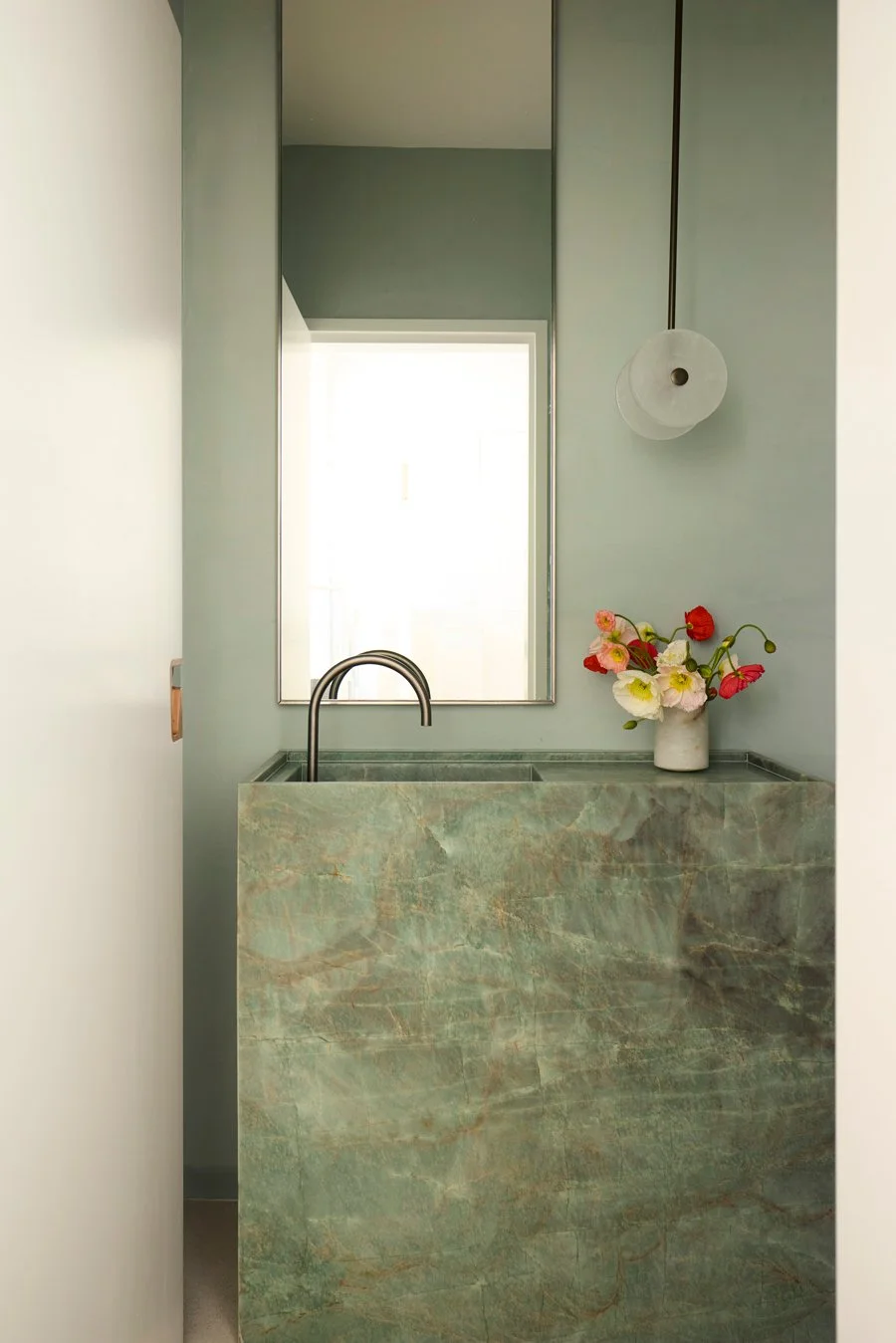Small bathroom with a green marble sink, a large mirror, a white ceramic vase with colorful flowers, and a white wall-mounted light fixture.
