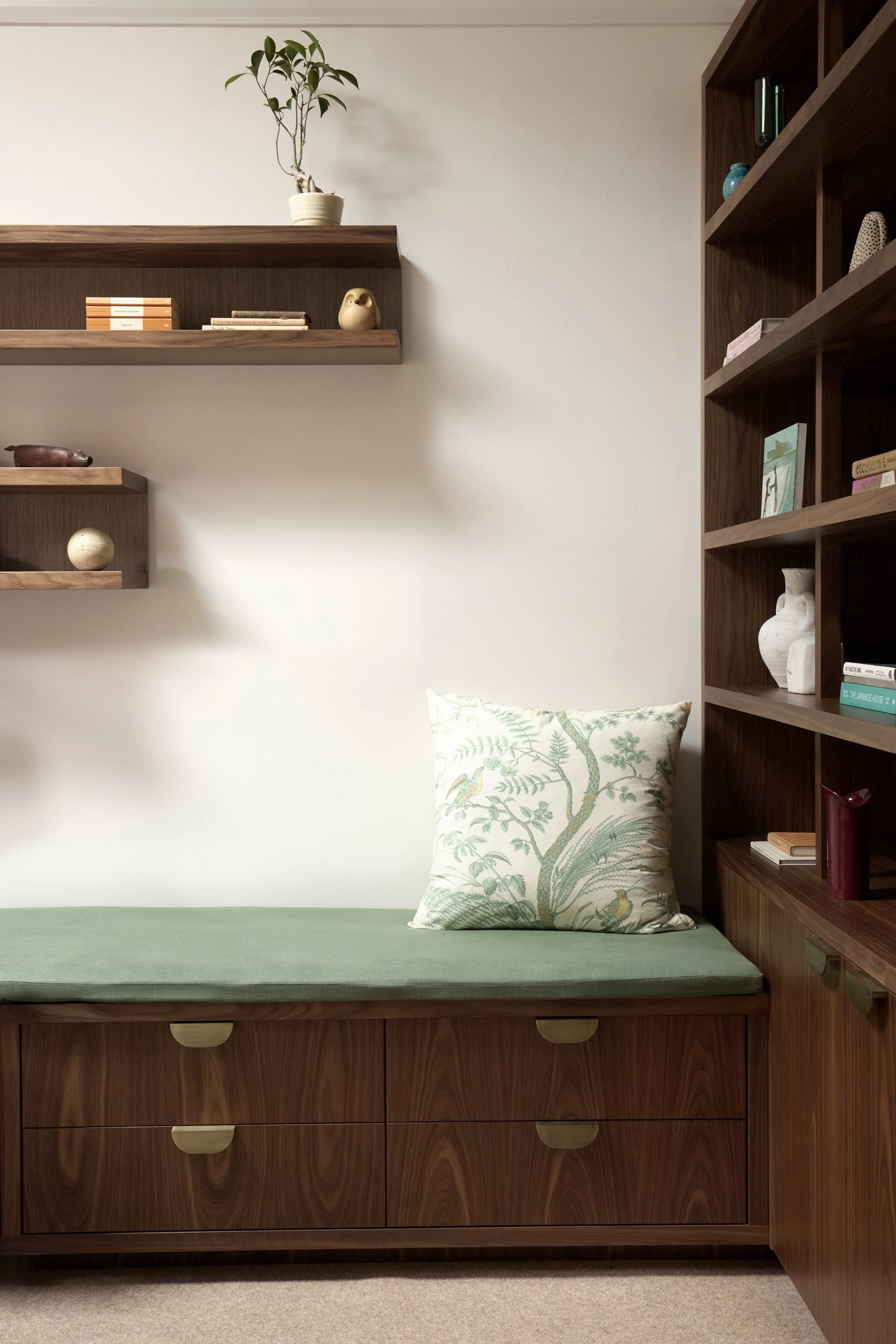 A room with wooden furniture, including a bed with a green mattress, a pillow with a bird illustration, and a bookcase with books and decorative items. The wall behind is plain, and there are two wooden shelves with decorative objects and books.