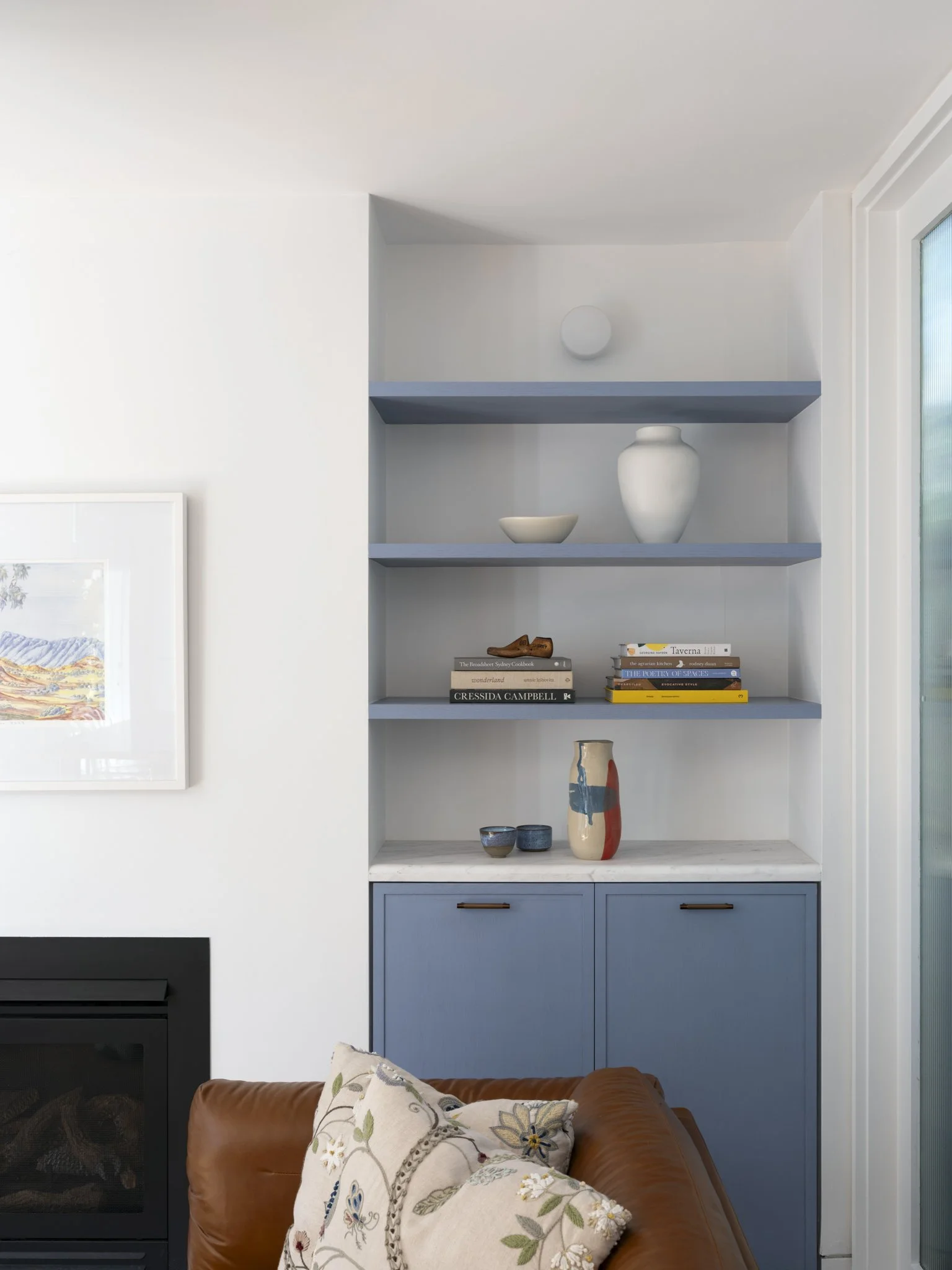 A built-in bookshelf painted in blue with decorative pottery and books, adjacent to a white wall and a living room with a leather sofa and a floral cushion.