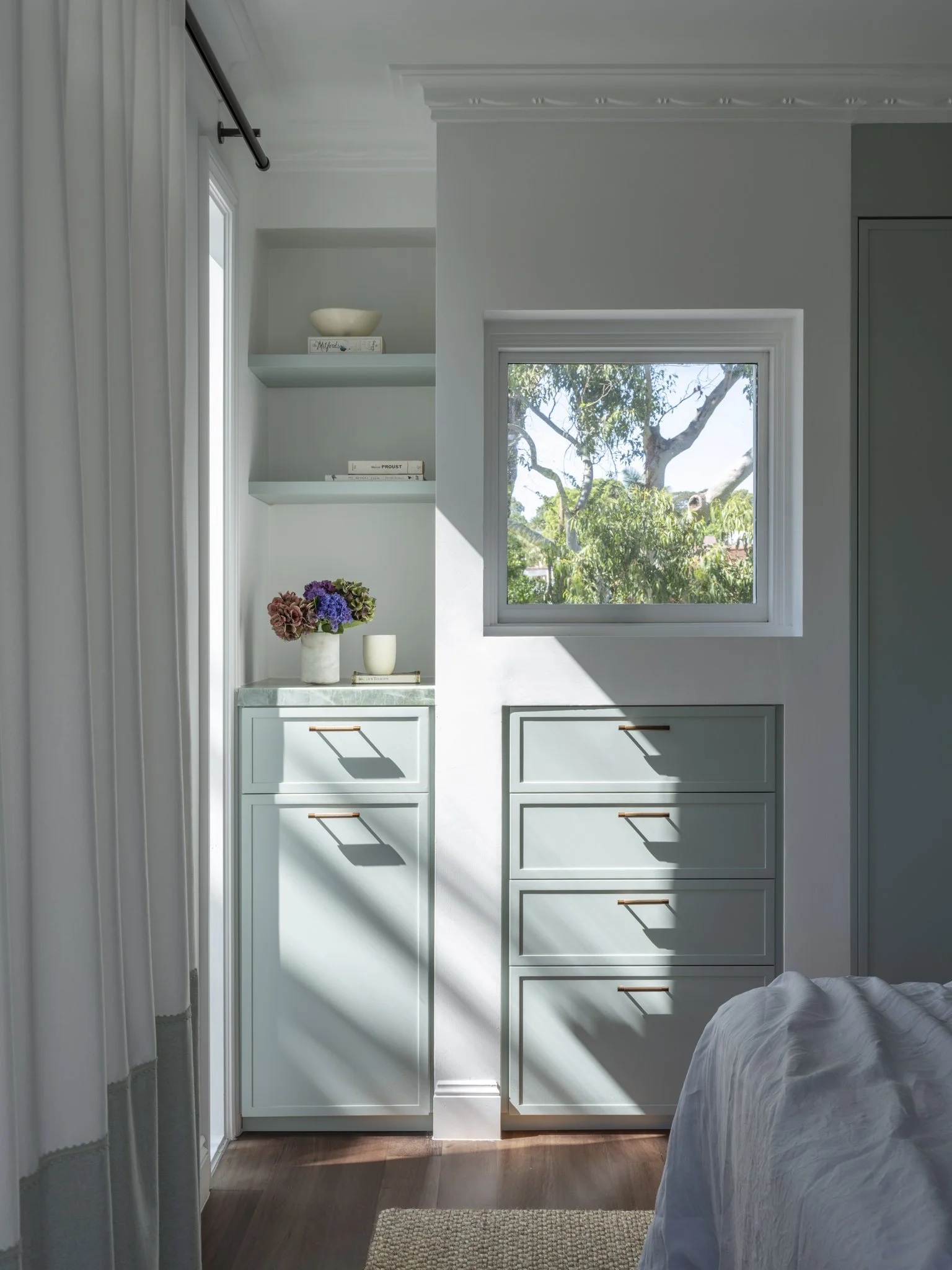 A bedroom with a window showing trees outside, a built-in shelf with books and decorative items, a flower arrangement on a small cabinet, and a bed with white bedding partially visible on the right side.