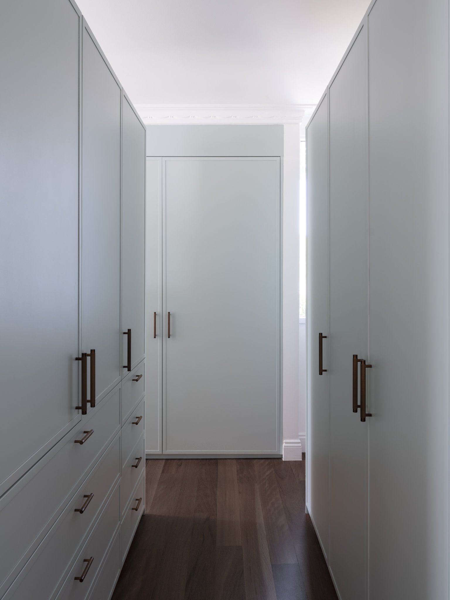 A walk-in closet with white cabinets and drawers, brown handles, and a dark wood floor, illuminated by natural light from a window at the far end.