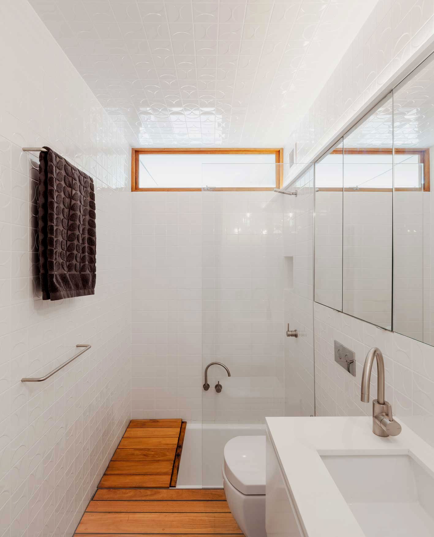 Modern bathroom with white tiled walls, a small window, a black towel on a towel rack, a wood mat on the floor, and a white toilet and sink.